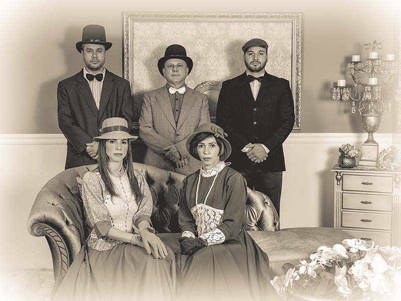 Retrato sépia de uma família de cinco pessoas em traje de época, três em pé e duas sentadas em sofá capitonê